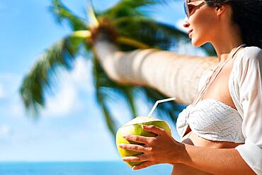 Beautiful young woman enjoy coconut drink and relax on tropical beach with palm tree. Summer vacation concept