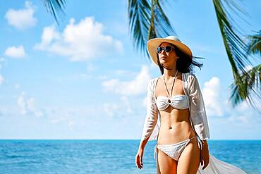 Pretty young woman in white bikini walking barefoot on the sea shore on tropical beach. Summer vacation and relax concept