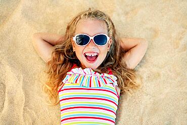 Top view of pretty little blonde girl in sunglasses lying on sandy beach during summer vacation. Sea relax, happy childhood concept