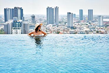 Young woman in hat enjoy summer vacation in roof top swimming pool with city view