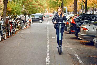 Young happy blond woman riding an electric scooter in the city, smiling at the camera, in autumn