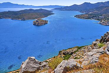 Aerial view of clear sea waters of harbor between Spinalonga island and peninsula, known as Kalydon, and Plaka village from above and rocky slope in foreground, Crete, Greece, Europe