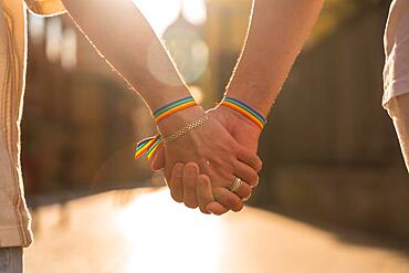 Detail of the holding hands of a couple of homosexual men with the rainbow flag at the pride party in the city at sunset, lgbt concept