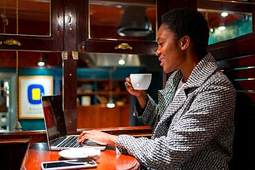 Business woman of African ethnicity in a coffee shop, working with a computer having a coffee