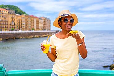 Black ethnic woman having an orange juice by the sea in the city, tourist enjoying the summer