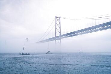 View of 25 de Abril Bridge famous tourist landmark of Lisbon connecting Lisboa and Almada in heavy fog mist wtih yacht boats passing under. Lisbon, Portugal, Europe