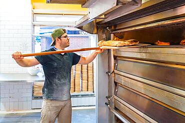 Baker of bakery in the workshop artisan workshop baking loaves in the oven, checking the baking