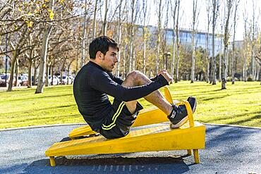 Confident young man doing abdominal exercises on a public equipment in outdoor fitness