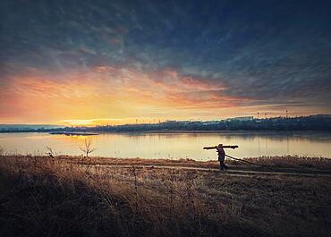 Person walking a country road carrying dry firewood. Peaceful sunset scene and wanderer silhouette on trail. Idyllic rural landscape