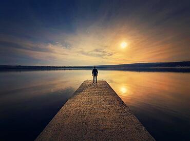 Man standing alone on the pier facing the sunset reflected in the calm lake water. Scenic view with a lonely person on the dock looking at the horizon