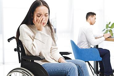 Worried young woman sitting wheel chair sitting front male colleague using laptop