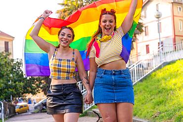 A closeup shot of two young Caucasian hugging females with LGBT pride flag outdoors