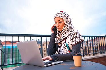 Arab girl with a white veil at the computer on the terrace of a coffee shop, with the computer and talking on the phone. Modern young man with new technologies