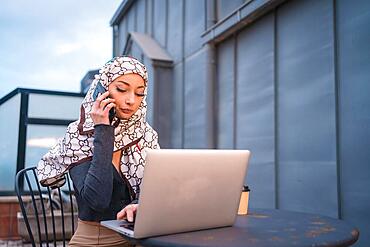 Arab girl with a white veil at the computer on the terrace of a coffee shop, with the computer and talking on the phone