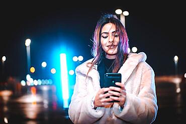 Caucasian brunette writing a message on the phone, wearing a pink wool jacket in an empty car park. Night urban session in the city