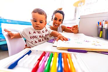 Young Caucasian mother playing with her in the room with toys. Baby less than a year learning the first lessons of her mother. Painting with color paints