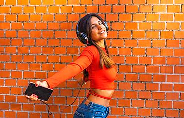 Lifestyle, young brunette with a wool cap and a red sweater with her eyes closed listening to music from her cell phone with her black headphones. Background of a brick wall