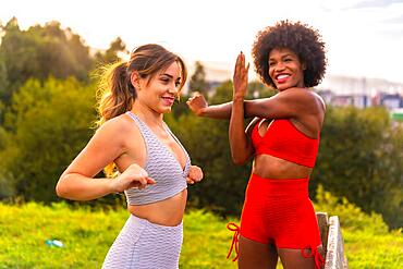 Caucasian blonde girl and dark-skinned girl with afro hair doing stretching before starting sports in the park. Healthy life, fitness, fitness girls, gray and red sport outfits