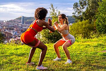 Caucasian blonde girl and dark-skinned girl with afro hair doing squat exercises in a park with the city in the background. Healthy life, fitness, fitness girls, gray and red sport outfits