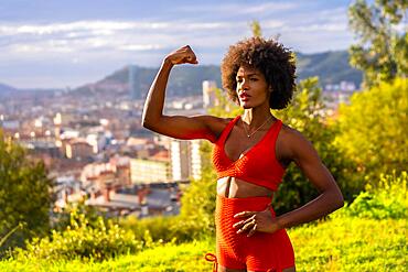 Fitness with a young black girl with afro hair, showing muscle, exercising in the field, red sport suit, fit girl, healthy life, copy paste space