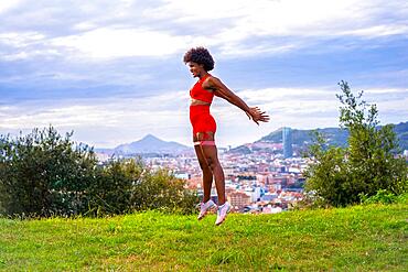 Fitness with a young black girl with afro hair, doing jumps and squats, exercising in the field, red sport suit, fit girl, healthy life, copy paste space