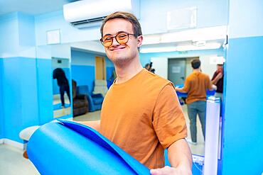 Happy and satisfied man with down syndrome after yoga class holing a mat