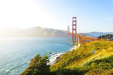 Sunlight provides high key highlights, lens flare over Marin Headlands with Golden Gate Bridge seen over rocky coastline at Fort Point during sunset in San Francisco, California. Horizontal copy space