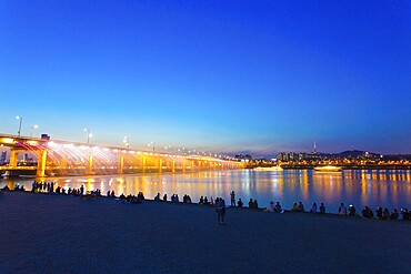 People watching Moonlight Rainbow water fountain and colourful light show from Banpo Bridge over the Han River at dusk on a clear night in Gangnam, Seoul, South Korea. Horizontal