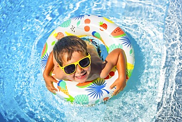 Top view of a cute boy in sunglasses playing in the swimming pool alone in summer time using rubber ring