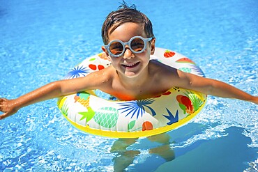 Cute boy with swim goggles and a rubber ring on a blue swimming pool smiling at camera