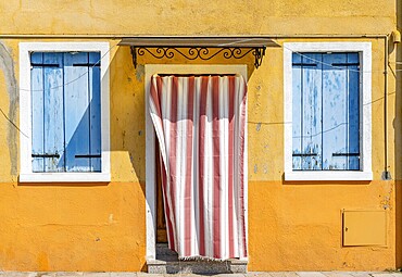 Yellow house facade with entrance door with red and white curtain and windows with blue shutters, colourful houses on the island of Burano, Venice, Veneto, Italy, Europe