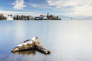 A peaceful lake with a log in the foreground with picturesque St George's Church on the lakeshore in front of snow-covered Pfänder on a clear winter's day, moated castle, Lake Constance, Bavaria, Germany, Europe