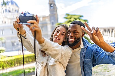 Multi-ethnic couple gesturing success while taking selfie with digital mirrorless camera in a park