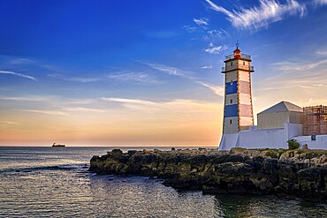Dramatic sunset on sea shore and Santa Marta lighthouse in Cascais, Portugal. Colorful sky, clouds, sunlight, low sun, calm ocean waters, local landmark, safety and navigation tower, rocky shore