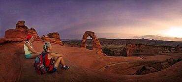 Hikers sitting at Delicate Arch natural stone arch at dusk, Arches-Nationalpark, near Moab, Utah, United States, North America