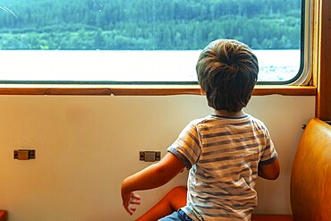 A young boy is sitting on a train window looking out at the water. The boy is wearing a striped shirt and shorts