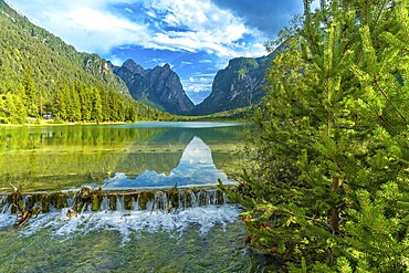 Crystal clear water cascading from a small waterfall into lake dobbiaco, reflecting the majestic dolomite peaks under a summer sky