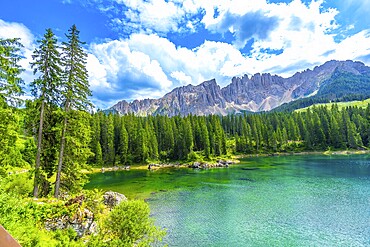 Emerald waters of lake carezza reflecting latemar mountain range, surrounded by lush pine forest under blue sky with white clouds in summer, dolomites, italy