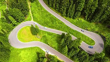 Aerial view of cars driving on a winding mountain road surrounded by lush green forest in the dolomites, italy, during summer