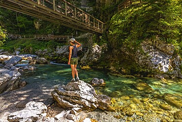 Female tourist standing on rock admiring tolmin gorges with wooden bridge, crystal clear turquoise water and lush vegetation in summer