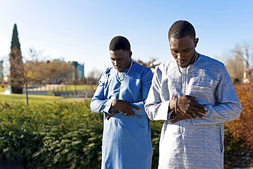 Two young senegalese muslim men wearing traditional dashiki clothing are performing salat prayer outdoors in a park during the holy month of ramadan