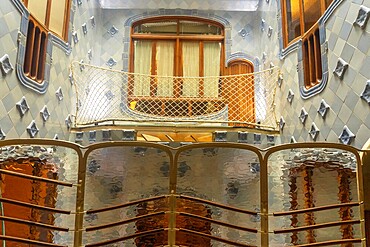 Light filtering through textured glass panels illuminates the unique blue tiled walls and wooden balcony of casa batllo, showcasing antoni gaudi's architectural brilliance