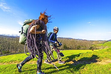 Two female climbers are walking across a mountain meadow carrying climbing rope and gear on a sunny day