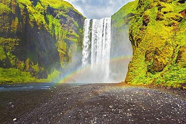 Picturesque huge rainbow appears in the water mist. The most popular waterfall in Iceland - Skogafoss. Water rushes down with a crash, forming a cloud of mist