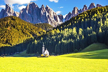 Dolomites, Tirol. Sunny autumn day. The famous symbol of the valley Val di Funes - church of Santa Maddalena. Rocky peaks and forested mountains surrounded by green Alpine meadows