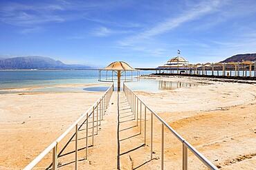 Railings enclose the path to the water. Lovely round umbrella protects from the sun. The drainless salt lake in the Middle East. Dead Sea. Coast of Israel