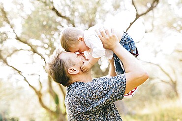 The father holds his son above him, they touching their noses, the son holds the father by the cheek .