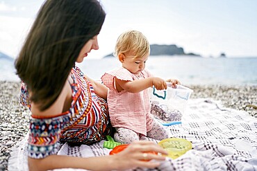 Mom lies on a blanket on a pebble beach next to a sitting little girl.