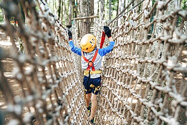 Boy walks across a mesh bridge, holding onto a cable. Back view.