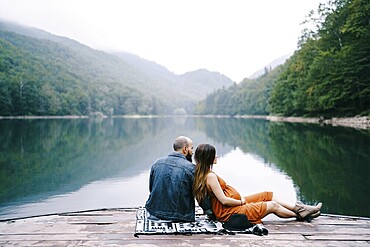 Man with pregnant woman are sitting on a pier by the lake in the park.